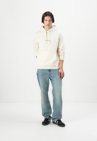 Licht crèmekleurige hoodie met een kangoeroezak, capuchon met trekkoord en geborduurd logo, gecombineerd met relaxte lichtblauwe jeans en zwarte sneakers.