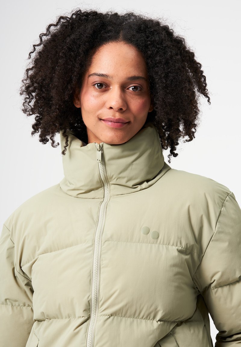Hellgrün gefütterte Jacke mit hohem Kragen, Reißverschluss und voluminösem, rautenförmigem Steppdesign. Verfügt über zwei dezente Punktakzente auf der Brust.