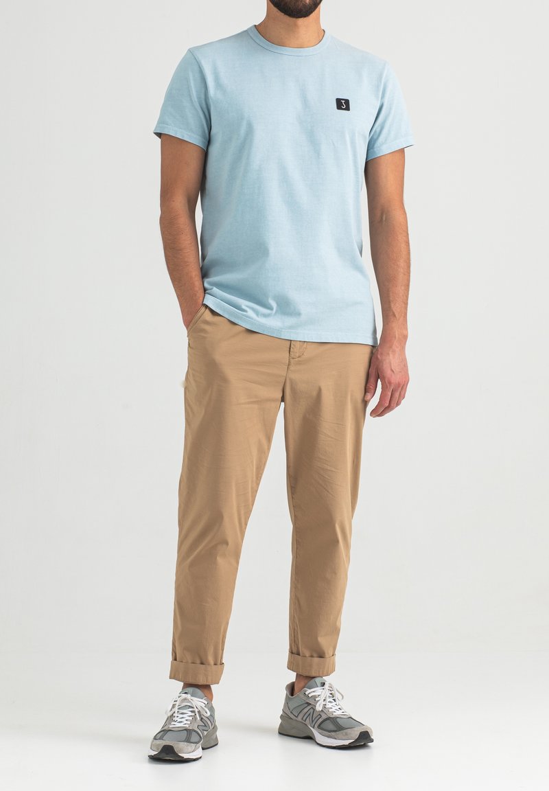 Helleblaue Baumwoll-T-Shirt mit Rundhalsausschnitt und kurzen Ärmeln, kombiniert mit beigen Chinos und grauen Sportschuhen. Schwarzes Logo auf der Brust.