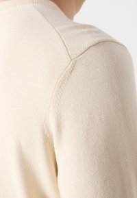 Gros plan d'une personne portant un pull en tricot beige, mettant l'accent sur les détails des coutures de l'épaule et du haut du bras.