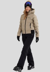 Puff jacket in licht beige met een strakke pasvorm, zwarte accenten en ritszakken, gecombineerd met zwarte broeken en zwarte handschoenen.