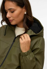 Wodoszczelna kurtka w kolorze oliwkowej zieleni z regulowanym kapturem, czarną podszewką z polaru, przednim zamkiem błyskawicznym oraz elastycznymi mankietami. Miękka, gładka faktura.