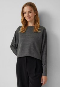 Femme aux cheveux blonds ondulés de longueur moyenne, portant un pull côtelé gris foncé et un pantalon noir, se tenant devant un mur gris clair.