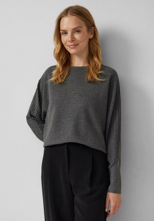 Femme aux cheveux blonds ondulés de longueur moyenne, portant un pull côtelé gris foncé et un pantalon noir, se tenant devant un mur gris clair.