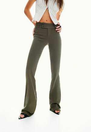 Pantalons évasés taille haute vert olive avec deux boutons et une texture lisse, associés à des chaussures à talons pointus noires.