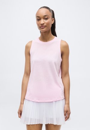 Femme avec queue de cheval foncée portant un haut sans manches rose clair et une jupe plissée blanche sur un fond clair uni.