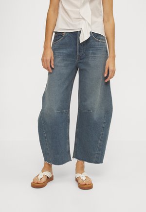 Person iført blå vide cropped jeans med frynset kant, hvide sandaler og en hvid bundet bluse på en ensfarvet hvid baggrund.