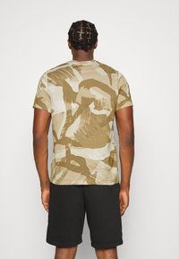 Camiseta de manga corta, ligera, en beige con patrones abstractos en marrón; con cuello redondo; combinada con pantalones cortos negros.