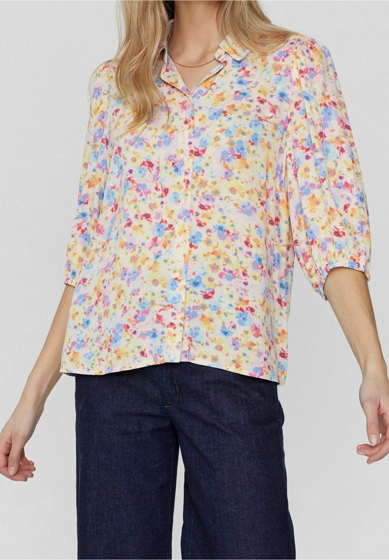 Chemise à boutons florale aux couleurs pastel, avec des manches bouffantes et un ourlet arrondi. Le tissu a une texture douce avec une sensation légère et aérienne.