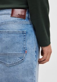 Jasnoniebieskie dżinsy z denimu z brązową skórzaną łatą na tylnej części pasa, wyposażone w małą metkę z flagą Union Jack na tylnej kieszeni.