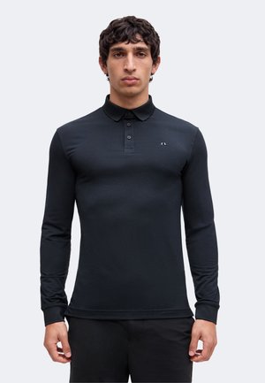 Mann mit kurzen lockigen Haaren, trägt ein eng anliegendes schwarzes Langarm-Poloshirt, steht vor einem schlichten hellen Hintergrund.