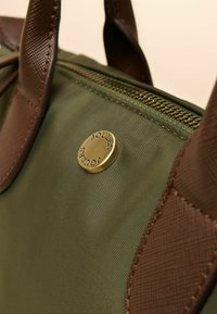 Zelená nylonová taška s hnedým textúrovaným lemom a mosadzným logom. Zapínanie na zips pozdĺž hornej časti. Hladký povrch bez viditeľných vzorov.