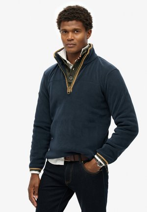 Marineblauwe fleece pullover met een hoge kraag, halve rits en een contrasterende tan rits. Gedragen over een gestructureerd grijs shirt en gecombineerd met donkere spijkerbroeken.