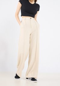 Pantalon beige à jambes larges avec plis doubles à l'avant, taille haute, poches latérales et fermeture à un seul bouton. Porté avec des baskets noires.
