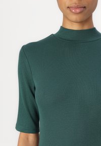 Haut du corps d'une personne portant un top côtelé vert foncé ajusté à manches courtes avec un col rond montant sur un fond uni.