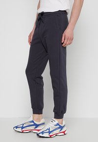 Donkergrijze sweatpants van zacht materiaal met een elastische tailleband, trekkoordsluiting en taps toelopende boorden. Gepaard met witte en blauwe sneakers.