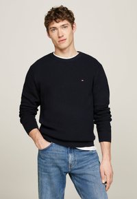 Maglione a maglia blu navy con texture a coste, colletto tondo e maniche lunghe. Piccolo logo decorativo sul petto; indossato con jeans azzurri.