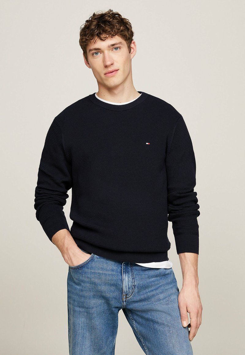 Maglione a maglia blu navy con texture a coste, colletto tondo e maniche lunghe. Piccolo logo decorativo sul petto; indossato con jeans azzurri.