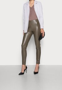 Frau in olivgrünen Lederhosen, mauvefarbener Bluse mit Knöpfen, helllavendelfarbenem Blazer, schwarzen High Heels, die eine schwarze Clutch vor einem weißen Hintergrund hält.
