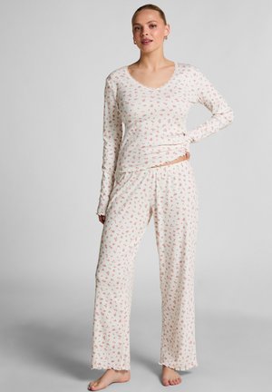 Woman standing barefoot wearing white long-sleeve top and pants with small pink floral print on a plain gray background.