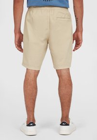 Beige bomullsshorts med elastisk midja, en enda bakficka och fållad finish, bärs med vita sneakers.