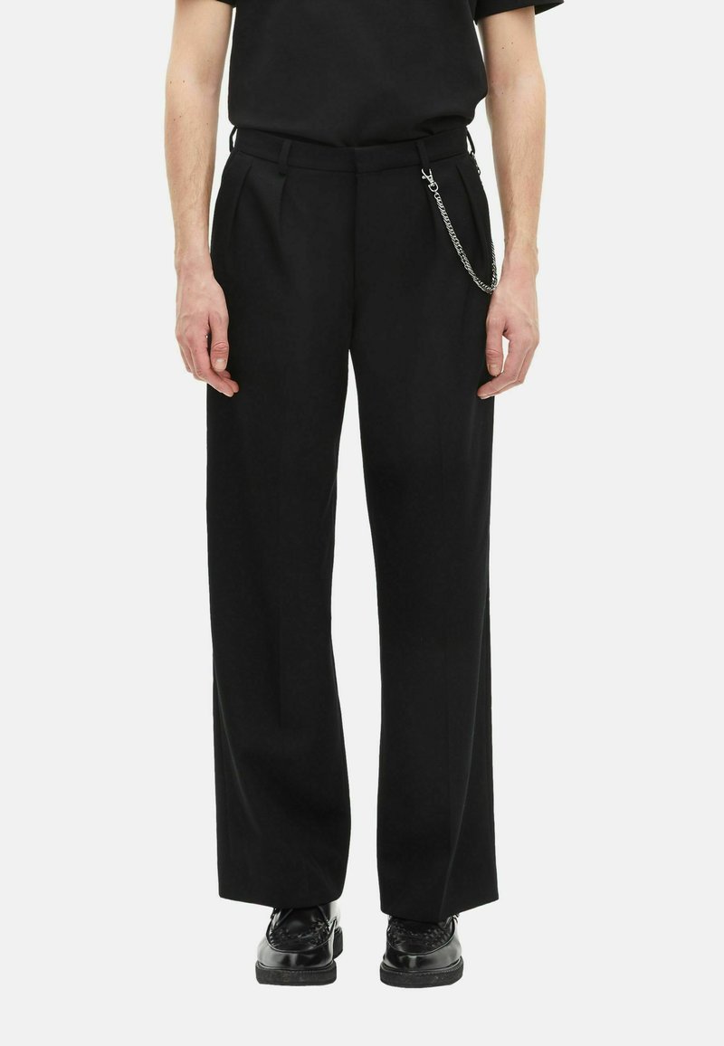 Hombre que lleva pantalones negros de pierna ancha con pliegues, un accesorio de cadena plateada, zapatos negros brillantes y una camisa negra.