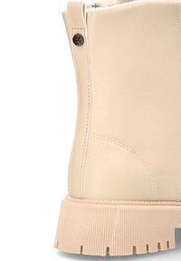 Botín beige con un upper sintético liso, suela de goma texturizada, costuras laterales y un detalle metálico circular cerca de la parte superior.