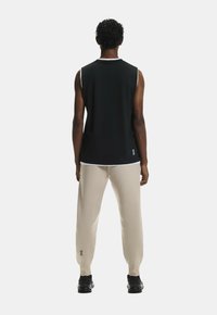 Schwarzes ärmelloses Oberteil mit weißen Akzenten, beiger Jogginghose und schwarzen Sneakers. Das Material wirkt glatt; die Passform ist lässig mit Logodetails an der Hose.