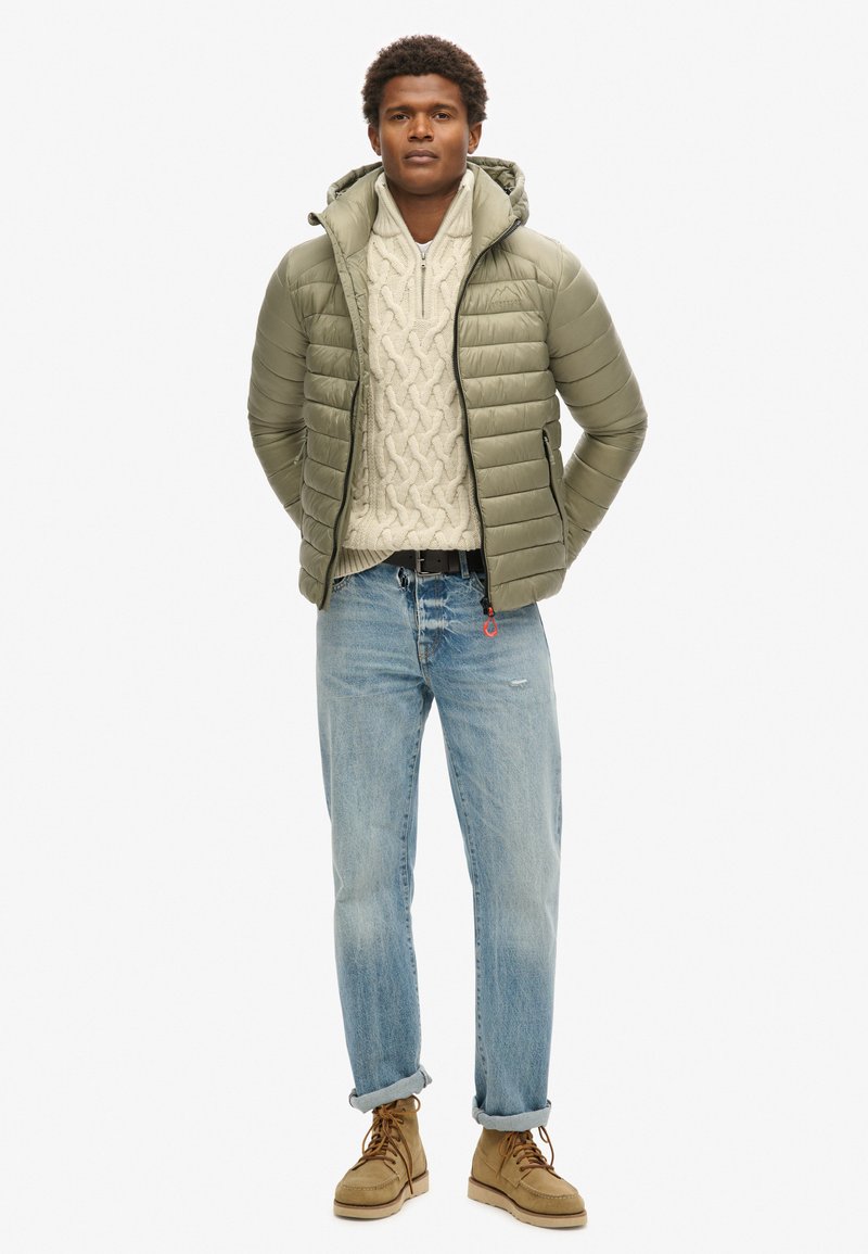 Mężczyzna stojący z rękoma za plecami, ubrany w oliwkową kurtkę puchową, kremowy sweter w sploty, jasnoniebieskie dżinsy i beżowe buty ze sznurówkami.