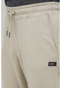 Lys beige sweathose lavet af blødt stof, med snoretræk i livet, sidelommer og et lille sort logo patch.
