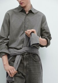 Grön rutig skjorta med knäppkrage, kombinerad med grå stickad tröja knuten runt midjan, bärs med grå jeans.