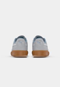 Un par de zapatillas Puma de ante gris con forro interior azul claro y suelas de goma marrón, mostradas desde la parte trasera sobre un fondo blanco.