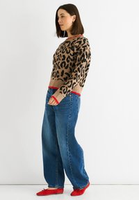 beige Pullover mit Leopardenmuster und roten Akzenten, kombiniert mit weiten blauen Jeans und roten flachen Schuhen, präsentiert in einer Seitenansicht.