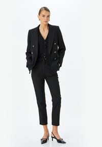 Blazer nero su misura doppiopetto con righe verticali, abbinato a un gilet aderente coordinato e pantaloni slim. Scarpe col tacco nere lucide.