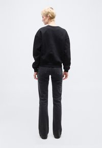 Άτομο με κοντά ξανθά μαλλιά που φοράει μαύρο φούτερ και σκούρο γκρι τζιν, στέκεται με την πλάτη προς τα πίσω σε ένα απλό φωτεινό φόντο.