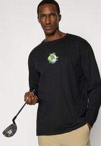 Μαύρο μακρυμάνικο μπλουζάκι με πράσινο κινούμενο χαρακτήρα και γραφικό με μπάλα γκολφ. Το μοντέλο κρατάει ένα γκολφ ρόπαλο, παρουσιάζοντας μια άνετη εφαρμογή.