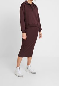 Persona vestida con una sudadera de cremallera color púrpura oscuro y una falda midi a juego, combinada con zapatillas altas blancas, de pie sobre un fondo liso.