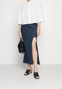 Bílá zkrácená košile na knoflíky, tmavá denimo sukně s vysokým rozparkem, černé lodičky na podpatku a černá taška typu bucket s detailním popruhem.