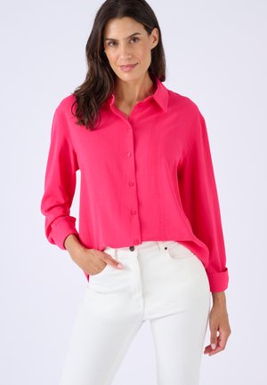 Femme aux cheveux foncés porte une chemise rose vif à manches longues boutonnée et un pantalon blanc, posant avec une main dans la poche devant un fond blanc.