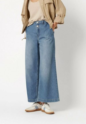 Frau trägt hellblaue Jeans mit weitem Bein, beiges Tanktop, braune Jacke und weiße Sneakers mit blauen Akzenten und steht vor weißem Hintergrund.