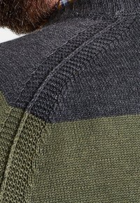 Pull en tricot avec trois sections : gris foncé en haut, vert olive en bas, et détails texturés le long de la couture de l'épaule.