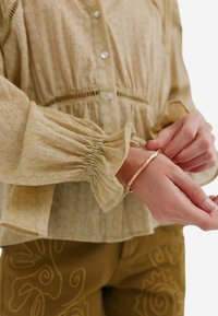 Blusa gialla chiaro, textured, con maniche a sbuffo e bottoni frontali, abbinata a pantaloni verdi oliva con motivo e un braccialetto dorato.