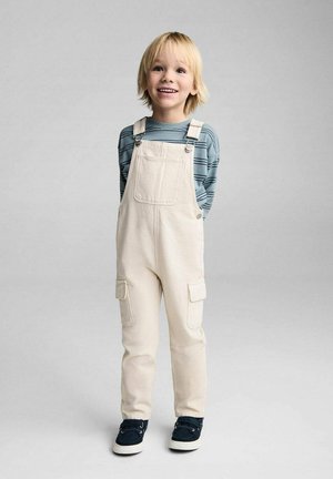 Salopette en coton blanche avec deux poches avant, portée par-dessus une chemise à manches longues rayée bleue. L'enfant porte des baskets noires. Fond lumineux.