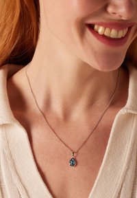 Collier en chaîne en argent avec un pendentif en forme de goutte d'eau en pierre précieuse bleue, monté dans un réglage délicat, porté sur un pull léger.