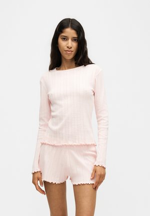 Jeune femme aux longs cheveux foncés portant un haut à manches longues côtelé rose clair et un short assorti avec des bords festonnés, debout devant un fond uni.