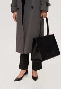 Black leather tote bag with embossed logo, carried by a person in a gray trench coat, black pants, and black pointed-toe heels.