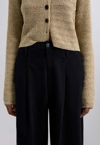 Cárdigan de punto texturizado beige con botones negros, combinado con pantalones negros de talle alto con pliegues y un cierre de botón en la cintura.