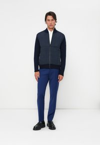 Chaqueta acolchada con cremallera en color marino, con mangas oscuras y diseño con cuello, combinada con pantalones azules y calzado negro, sobre un fondo blanco.