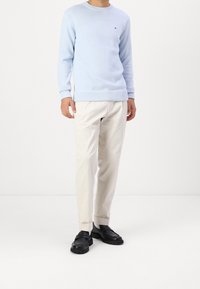 Pull en maille bleu clair avec un col rond, associé à un pantalon beige et des chaussures noires à enfiler. Le pull présente un petit logo sur la poitrine.
