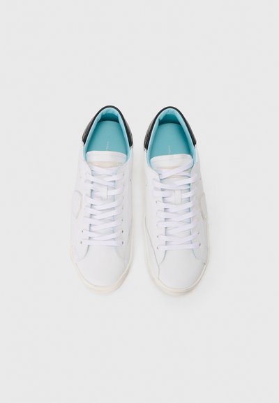 Baskets blanches à la texture lisse, avec un capuchon de talon noir, une doublure bleu clair et des lacets blancs. Design minimaliste avec des accents subtils.